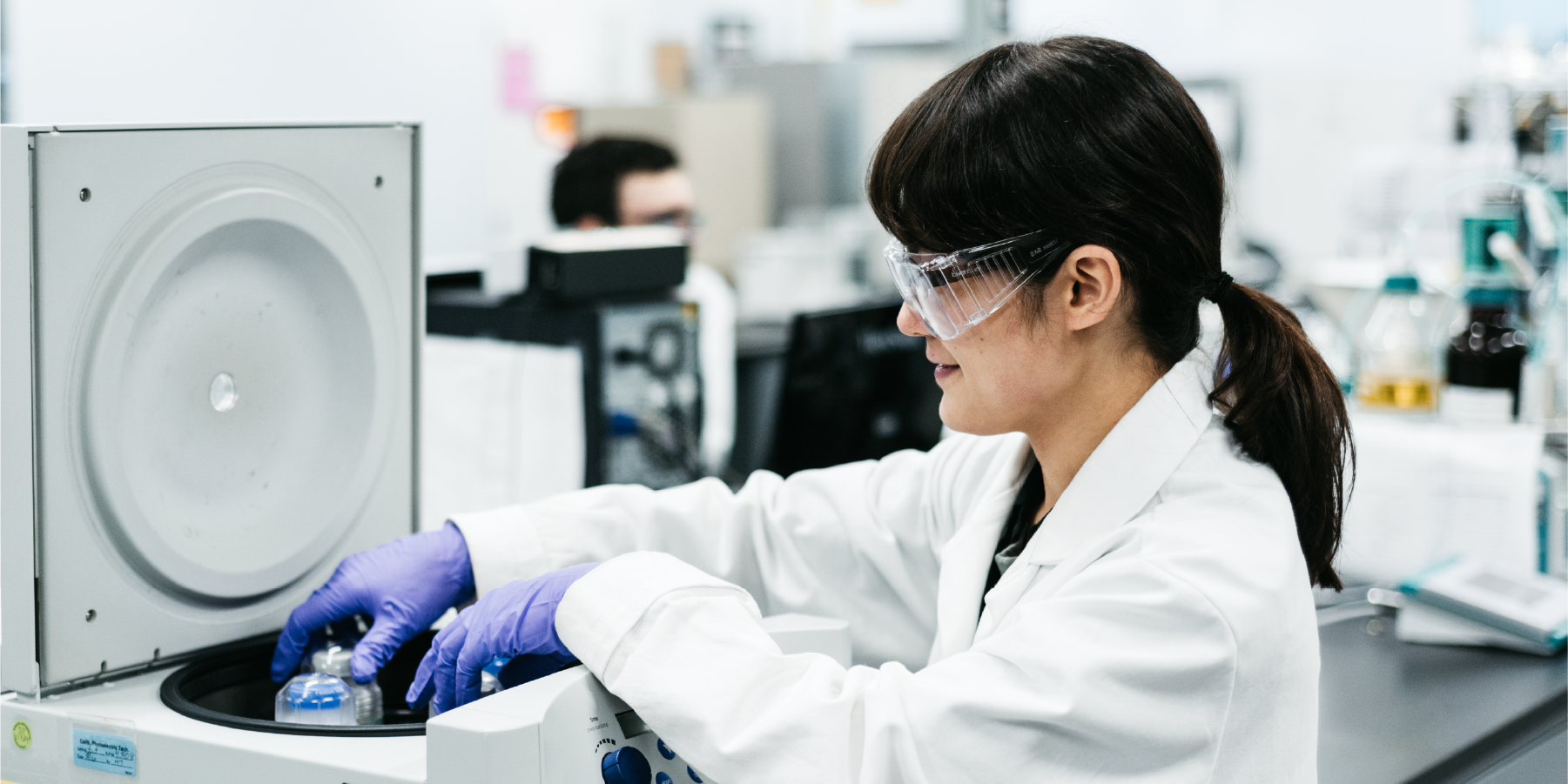 Woman in lab coat and goggles looking at beaker of lotion being stirred in a laboratory